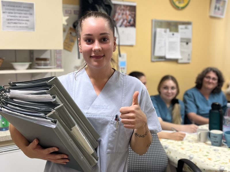 Weibliche Auszubildende hält Patientenakten im Arm und zeigt den hochgestreckten Daumen