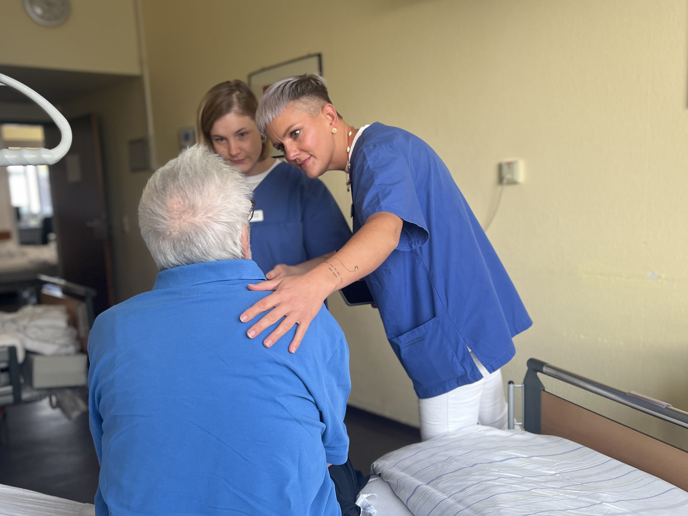 Zwei Pflegefachfrauen sprechen mit einem Patienten, der im Krankenhausbett sitzt
